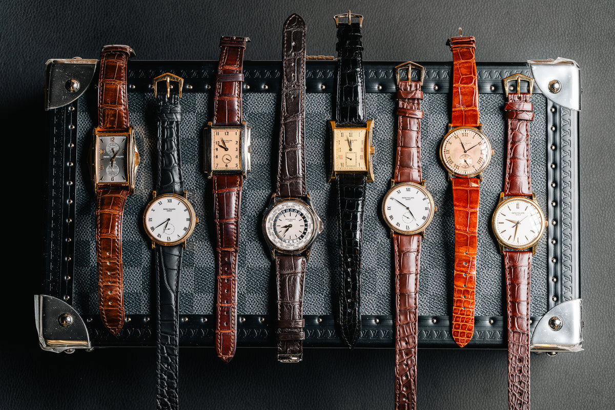 Eight vintage Patek Philippe watches on leather straps draped over a studded travel trunk